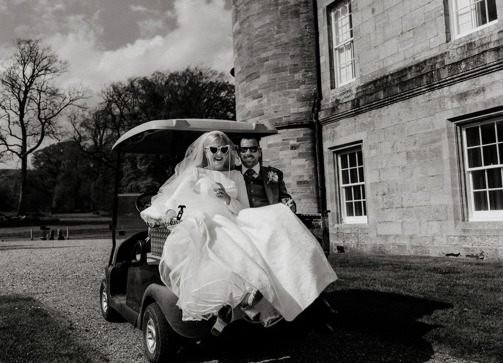 A couple rides in a golf cart outside a stone building on their wedding day.