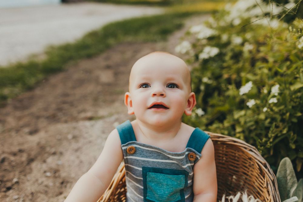 Smiling baby in teal overalls poses in wicker basket by garden path.