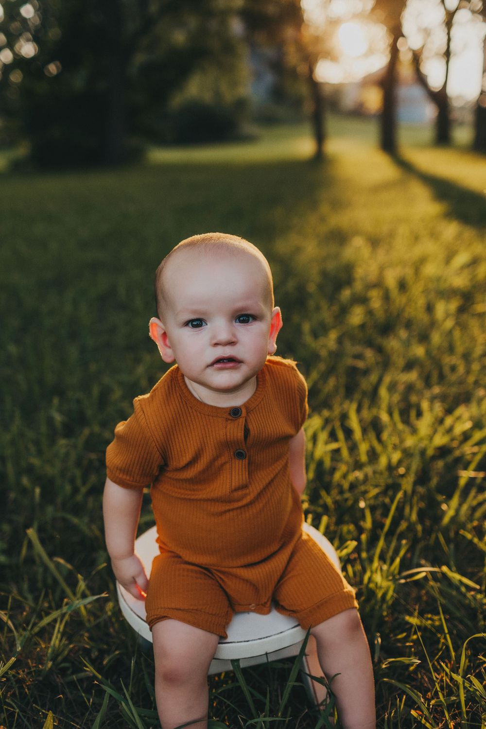 Baby wearing orange outfit sits outdoors in evening sunlight.