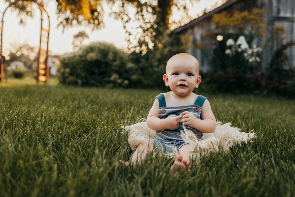 A little one sits on a blanket in the grass wearing blue overalls during golden hour.
