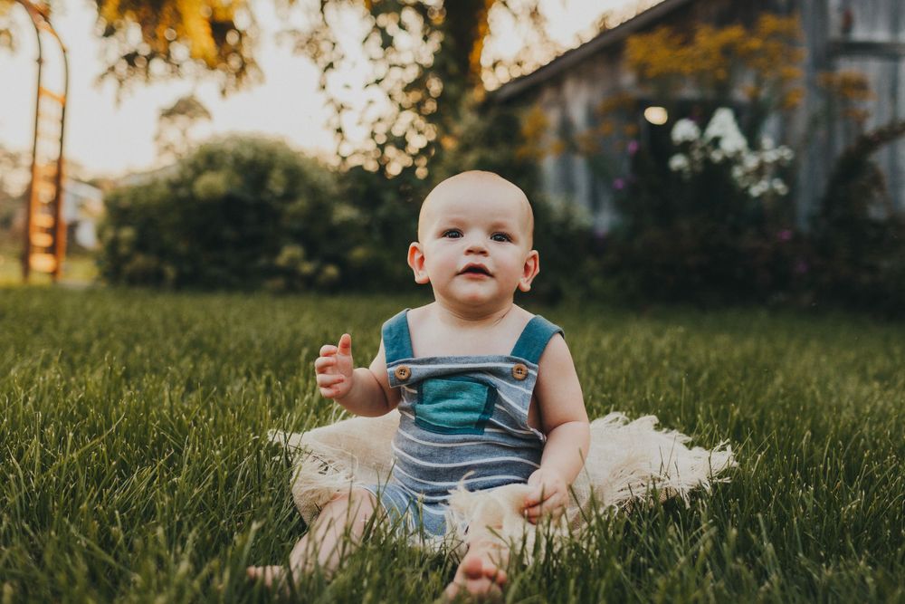 Baby in teal overalls sits on blanket in evening sunlit yard.