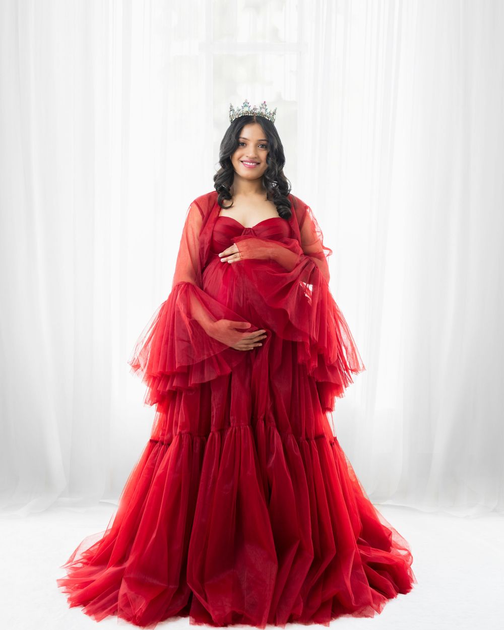 Dramatic maternity portrait in a flowing red tulle gown and crown against white backdrop.