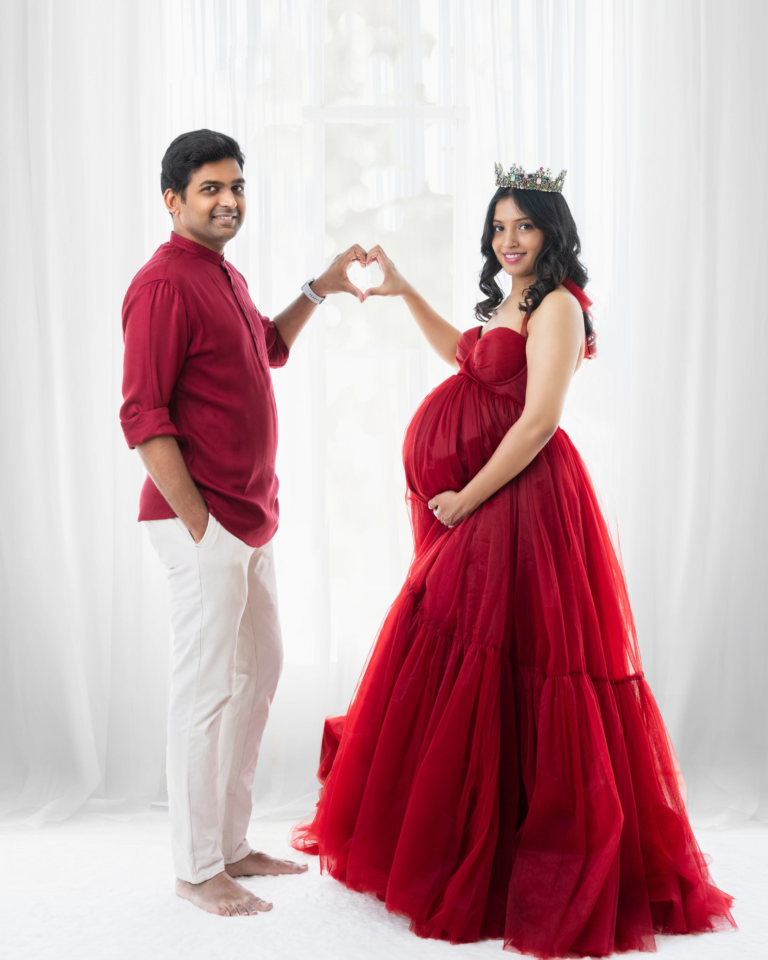 Couple forming heart shape with hands during maternity photoshoot in red and white outfits.
