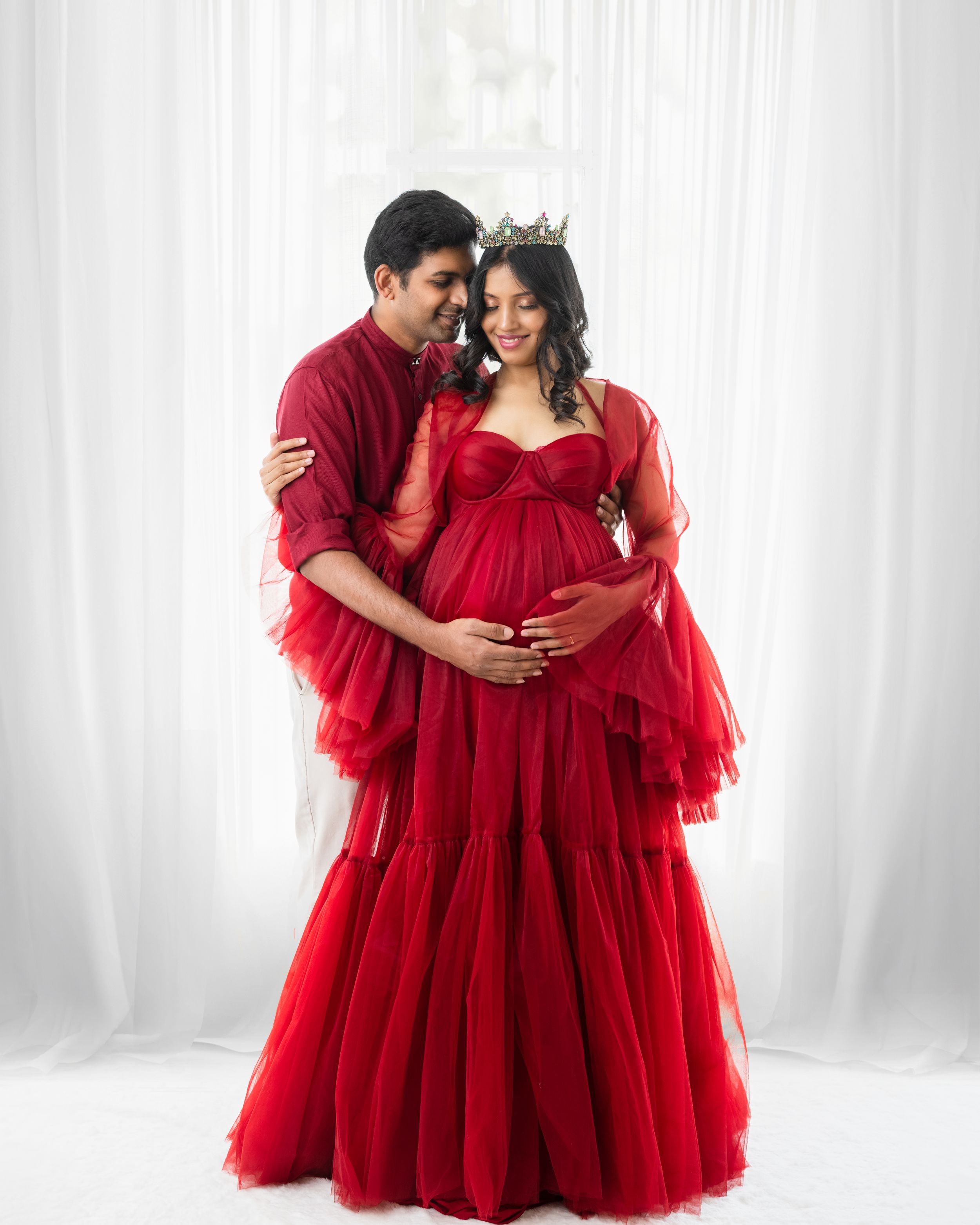 Intimate maternity portrait of couple embracing in matching red outfits against white background.