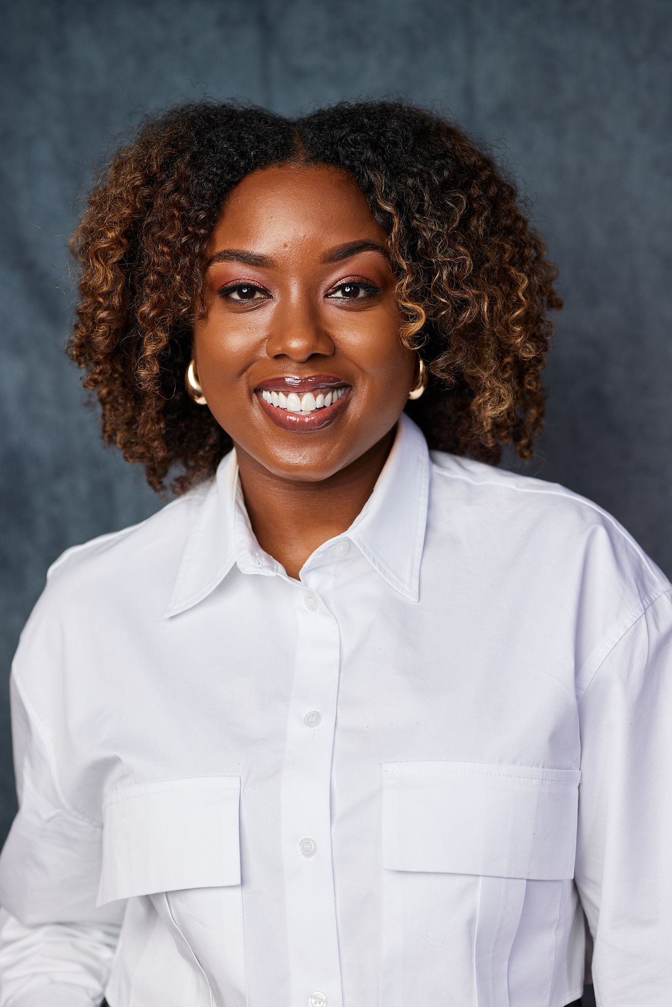 Confident young Black woman smiling in white button-up shirt against dark gray background.