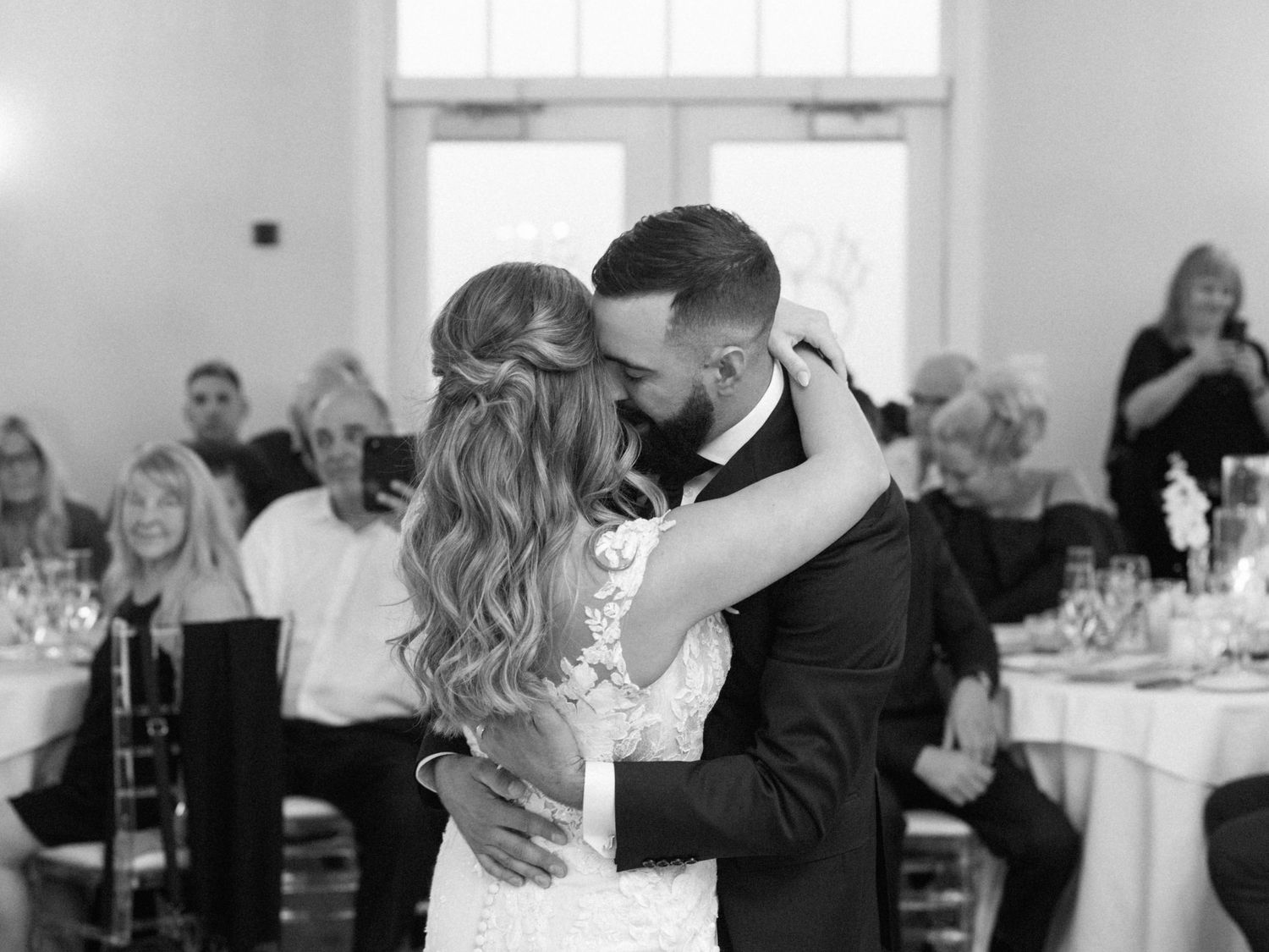 A groom leans down to his bride's ear, arms wrapped closely around each other as they dance at their reception.