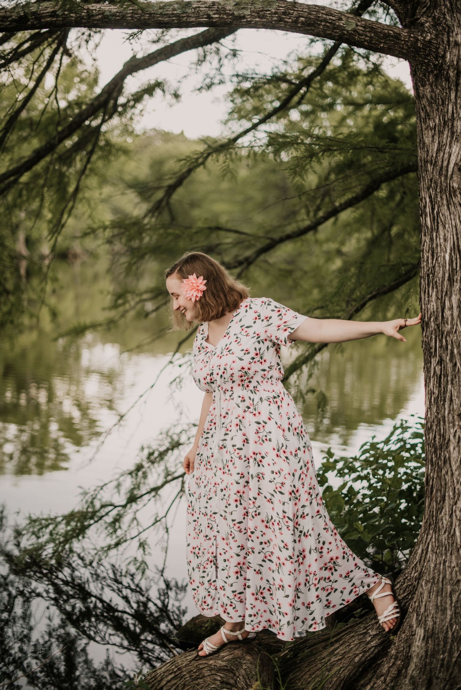Person in a floral dress twirls gracefully by a serene lake beneath overhanging trees.