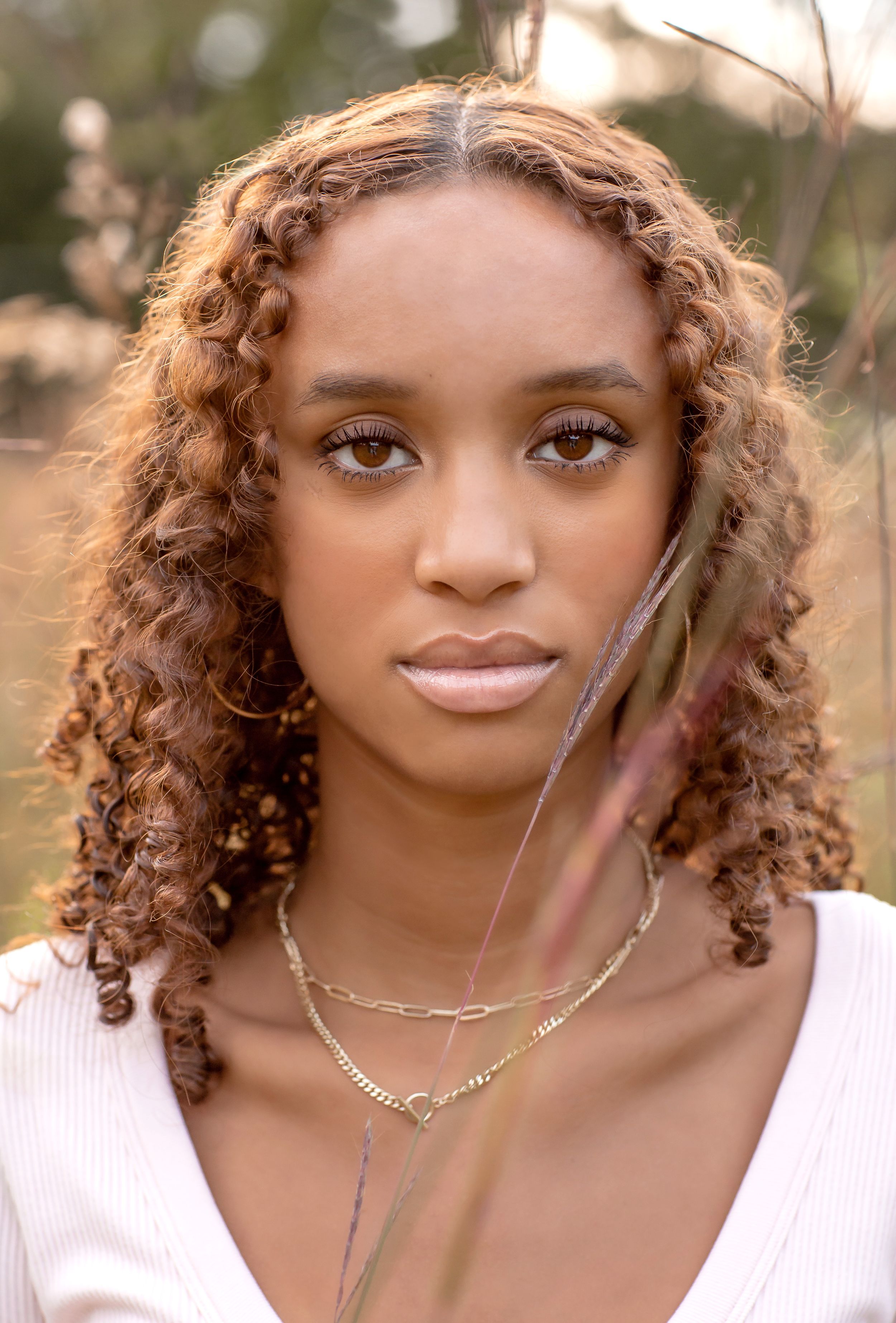 Young woman with curly auburn hair and gold layered necklaces poses outdoors in a white top.