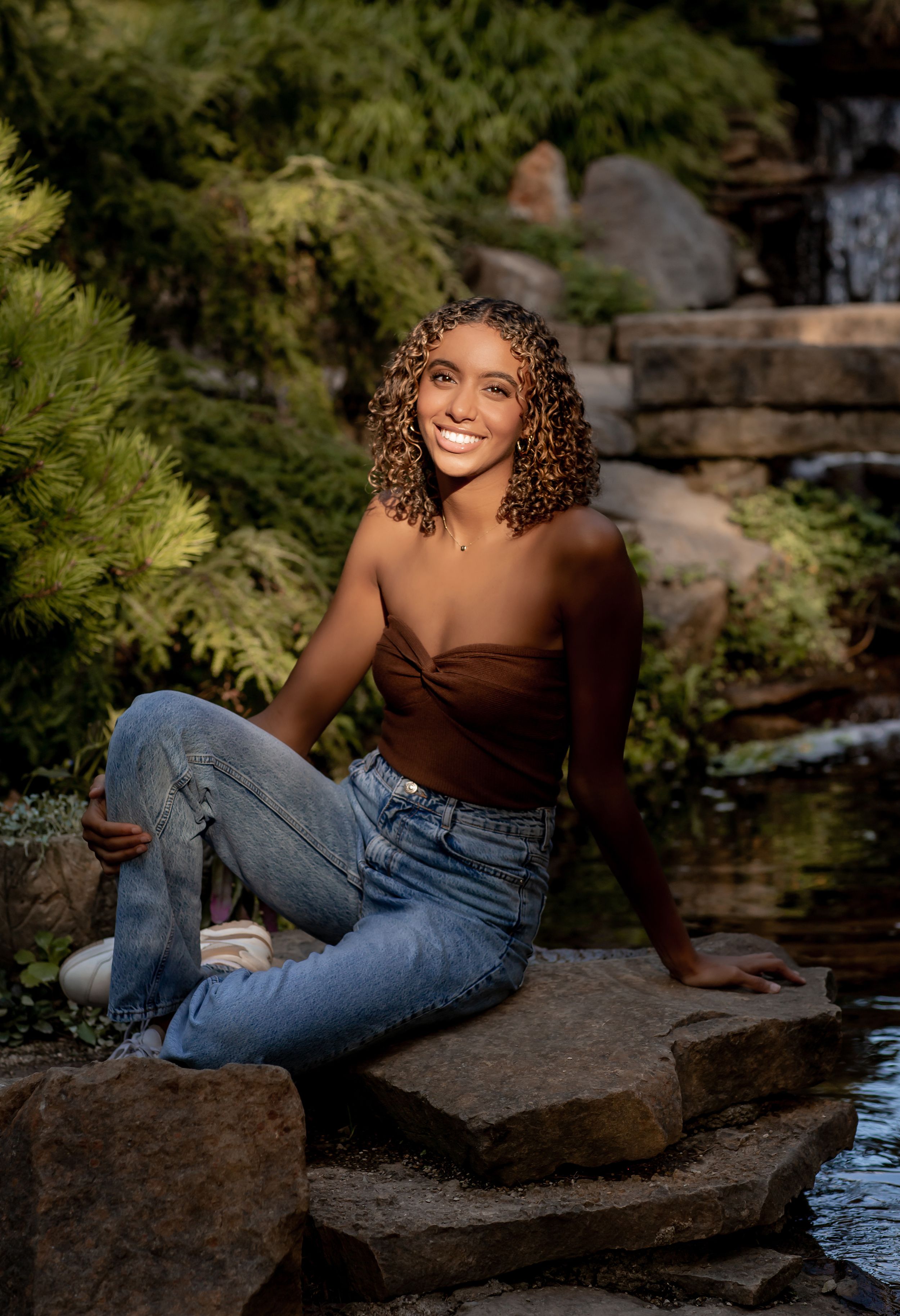 Smiling young woman with curly hair sits on rocks in a brown tube top and jeans near a waterfall.