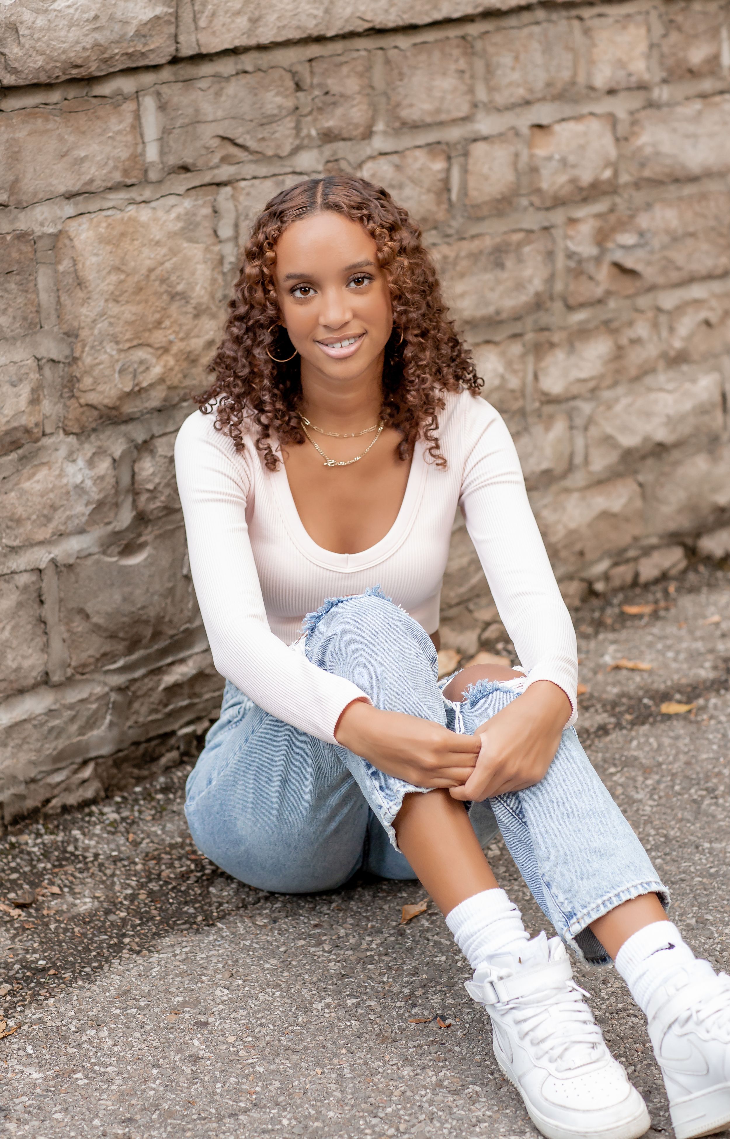 Smiling young woman with curly hair sits against a stone wall in a white top and ripped jeans.