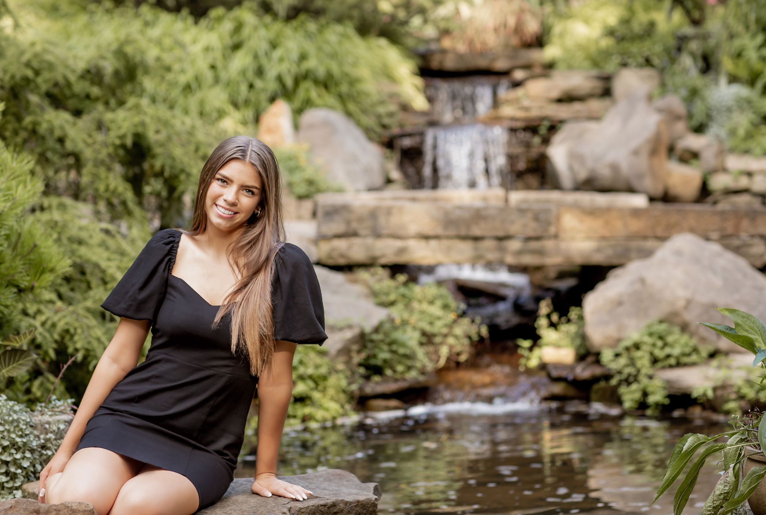 A young woman in a black dress sits on rocks beside a serene waterfall in a lush garden.