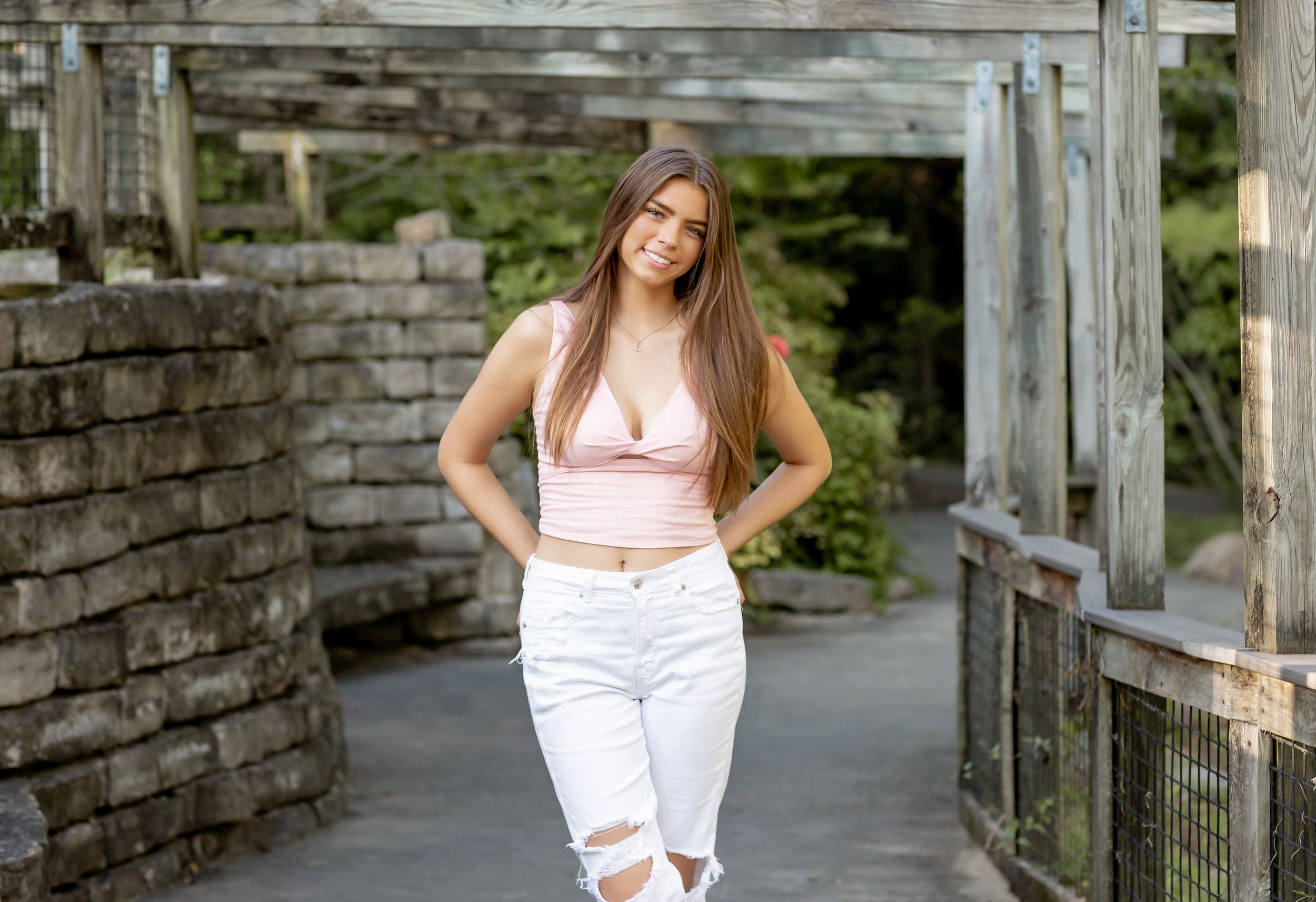 A smiling young woman in a pink top and white ripped jeans poses under a rustic wooden structure.
