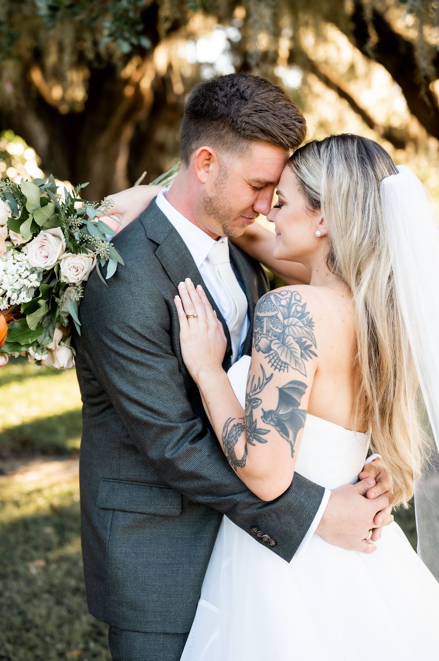Bride with floral tattoos and groom share tender moment during outdoor wedding photoshoot.
