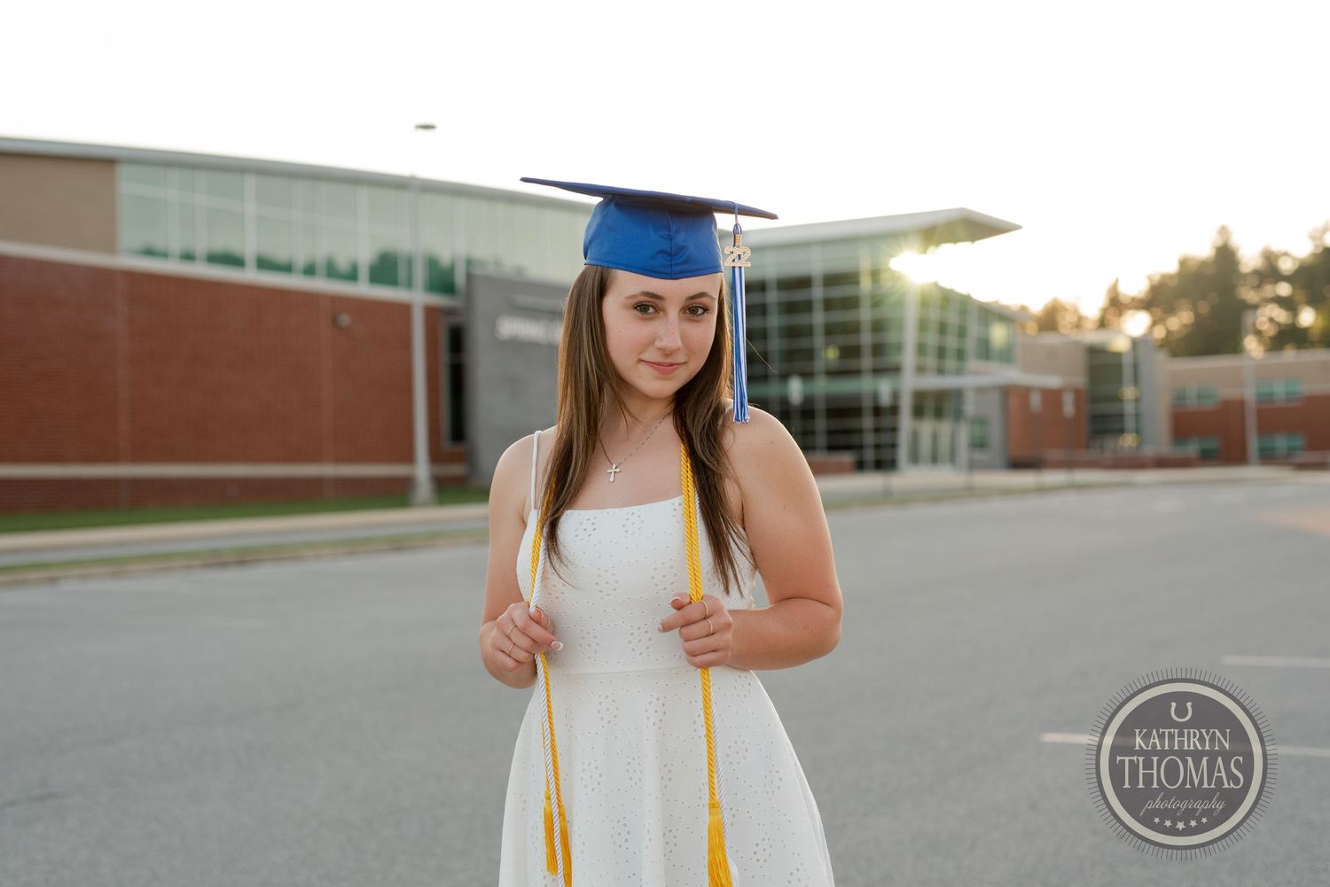 Graduate poses in white dress and blue graduation cap with gold tassel outdoors.
