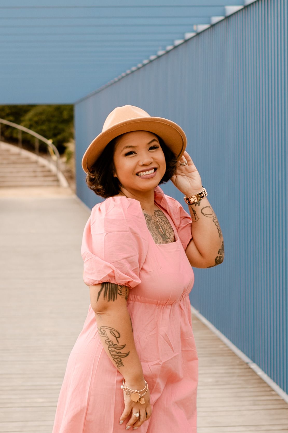 A person in a pink dress and tan hat poses confidently against a blue wall outdoors.