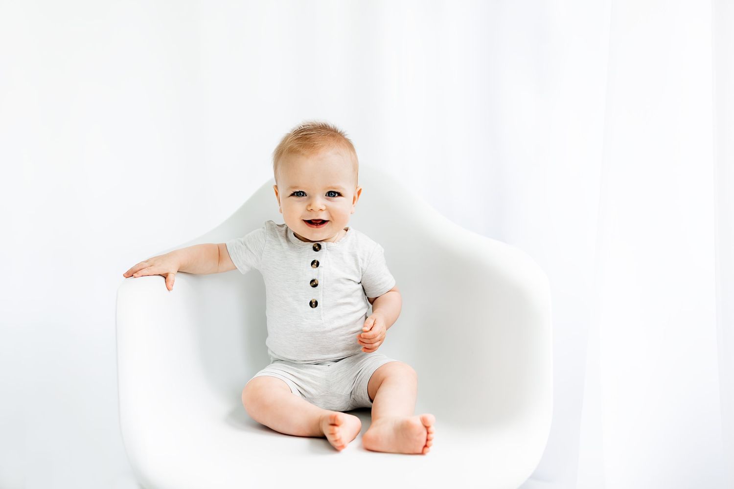 Baby wearing white onesie sitting and smiling in modern white chair.