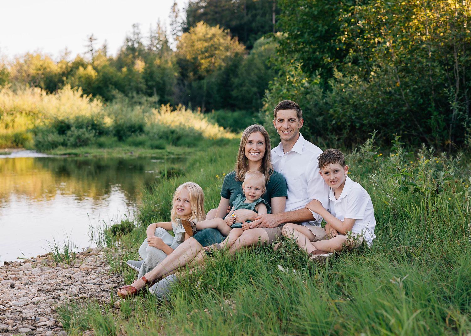 Family sitting together on grass near a peaceful pond during golden hour.