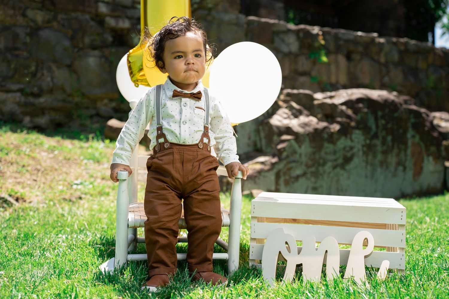 A young child in suspenders and bow tie poses near white furniture one year old shoot