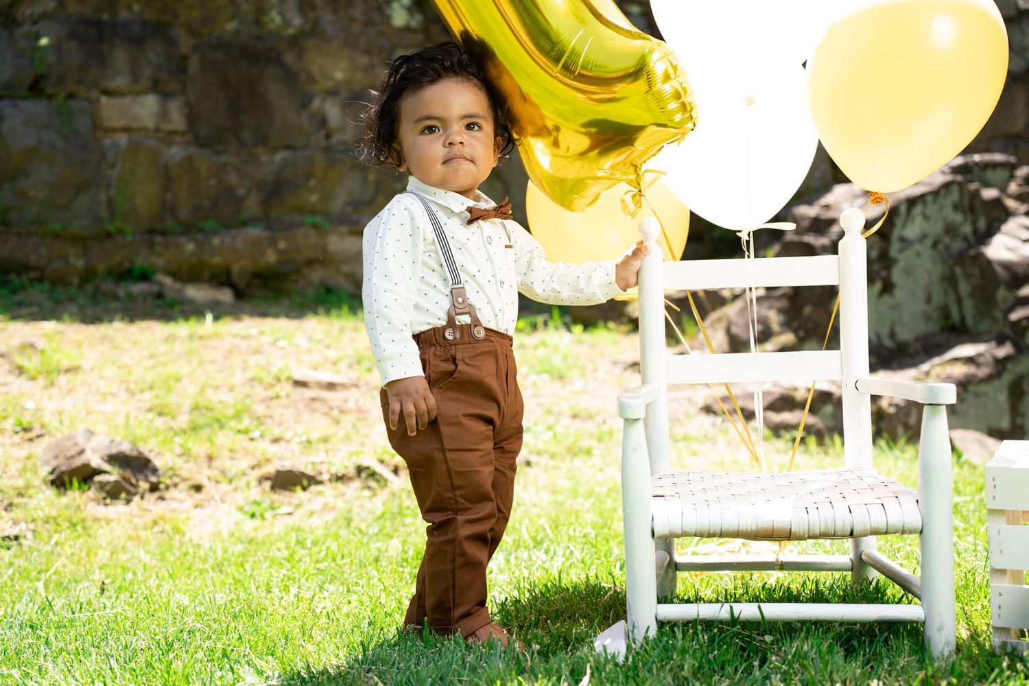 A toddler in brown overalls and white shirt stands on grass near white chairs and balloons