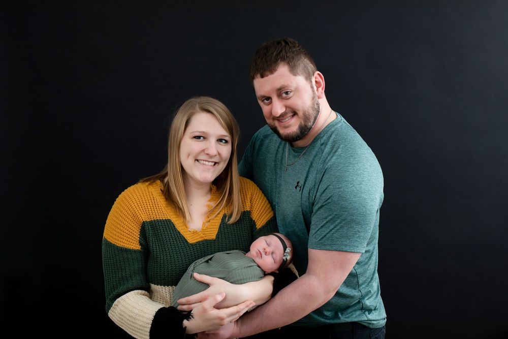Family wearing green and yellow sweaters poses with newborn against dark backdrop.