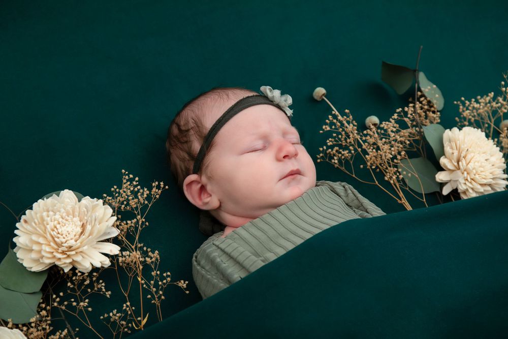 Sleeping infant in green wrap with white flowers and dried botanicals on dark green background.