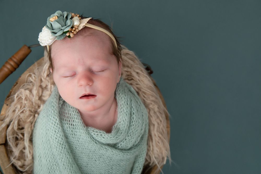 Newborn in mint green wrap sleeping peacefully in wooden bowl against grey background.