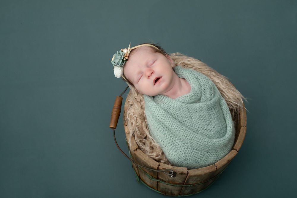 Baby wrapped in mint swaddle sleeping in rustic wooden bowl on teal backdrop.