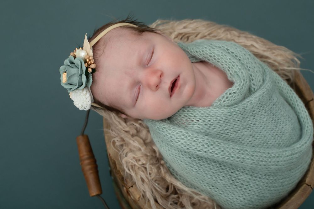 Sleeping newborn swaddled in mint knit wrap with floral accent in wooden basket.