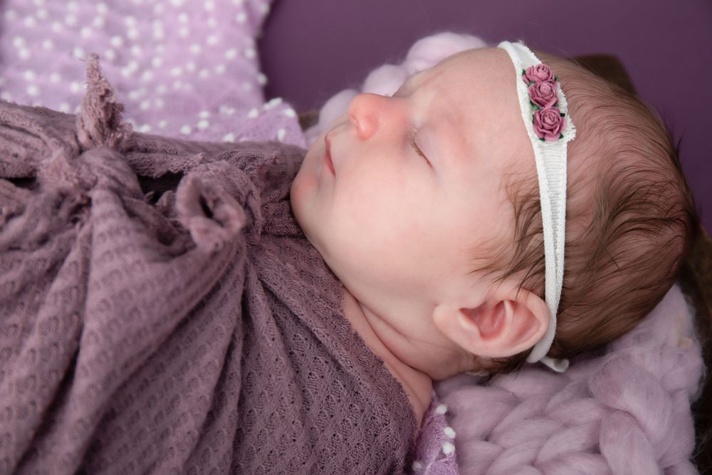 Peaceful sleeping infant wrapped in mauve fabric on purple dotted background.