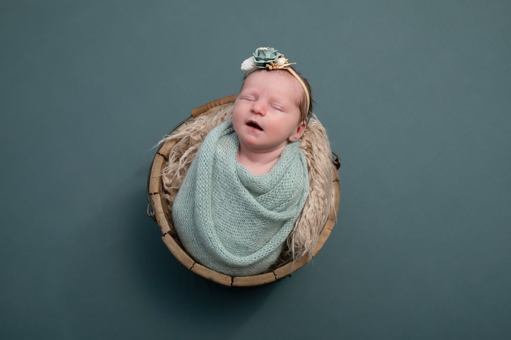 Sleeping baby swaddled in mint wrap nestled in wooden bowl against grey backdrop.