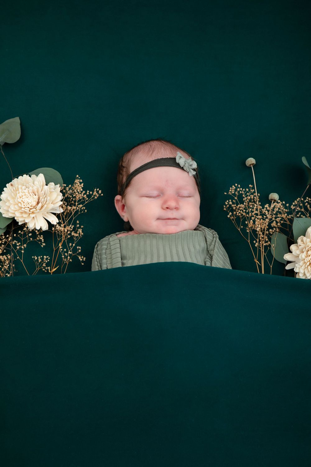 Newborn wrapped in sage green swaddle lying on dark green backdrop with white flowers and dried baby's breath.