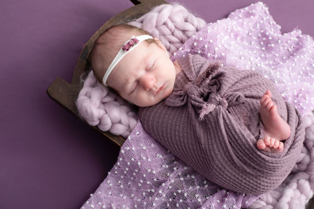 Infant in grey wrap sleeping on purple dotted fabric with delicate lace details.