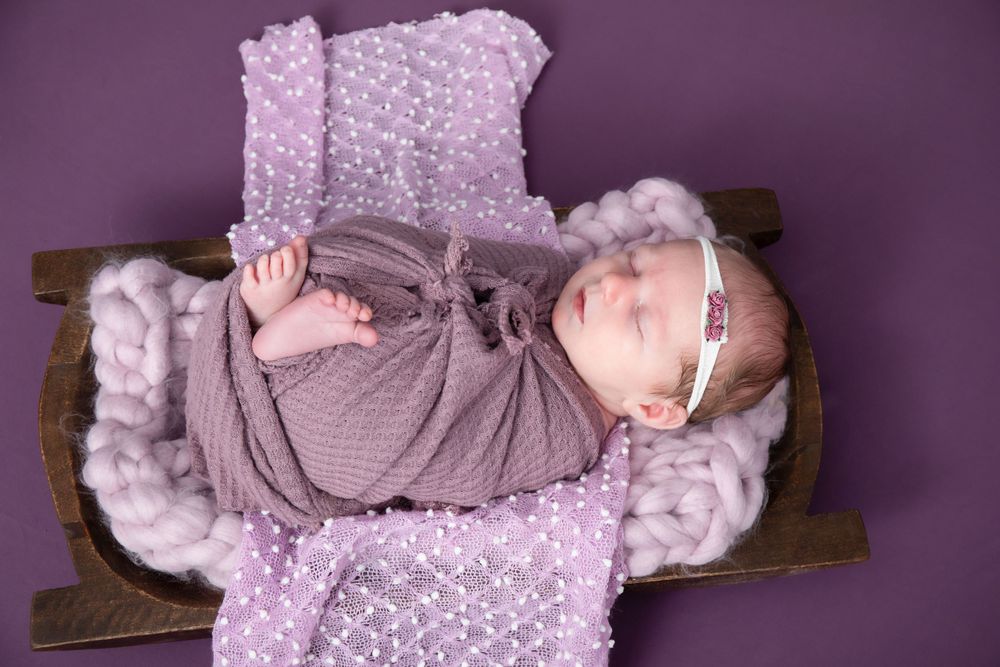 Sleeping baby in grey wrap on purple textured blanket surrounded by soft textures.