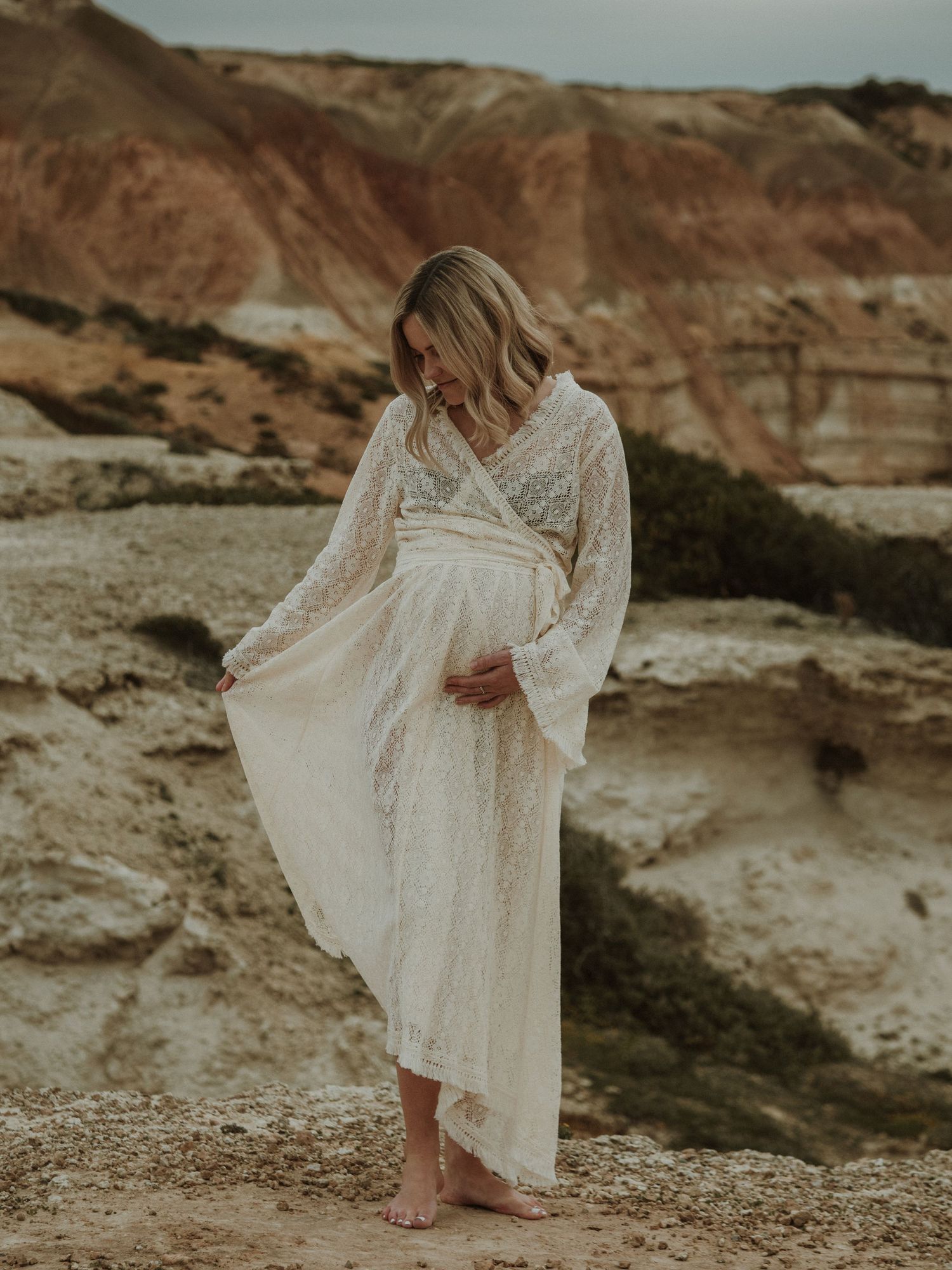 Ethereal maternity photo with flowing white dress against rocky coastal landscape.