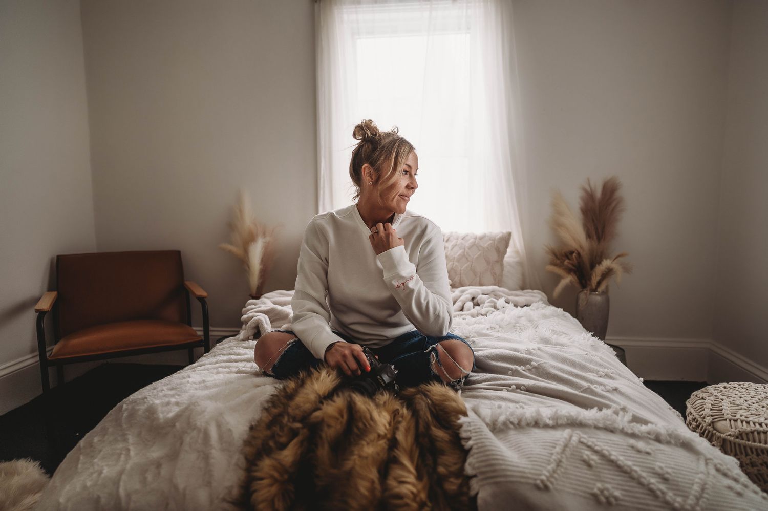 Person sits thoughtfully on bed with dried pampas grass decor in bright minimalist room.