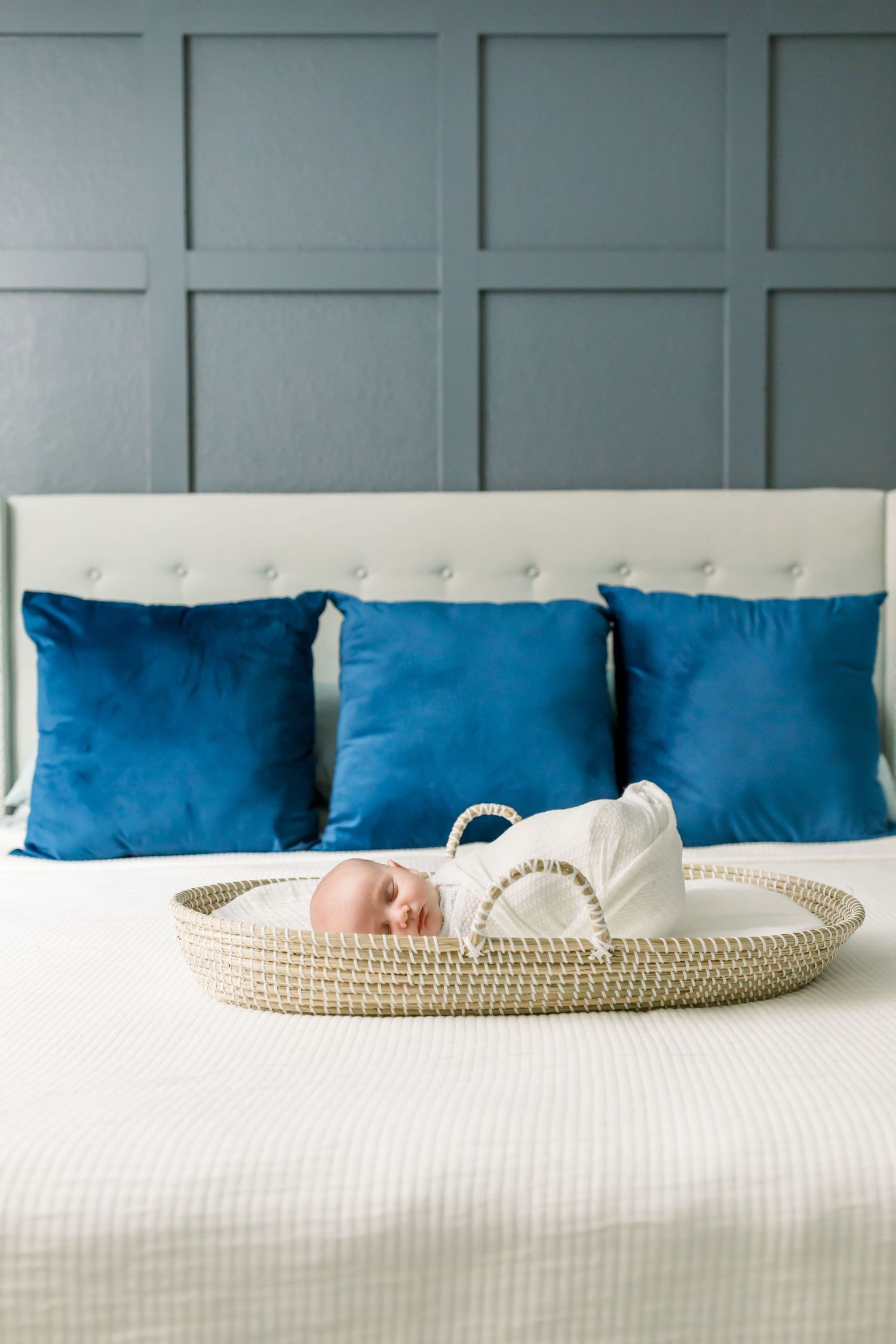A sleeping newborn rests in a woven basket on a white bed with vibrant blue throw pillows.