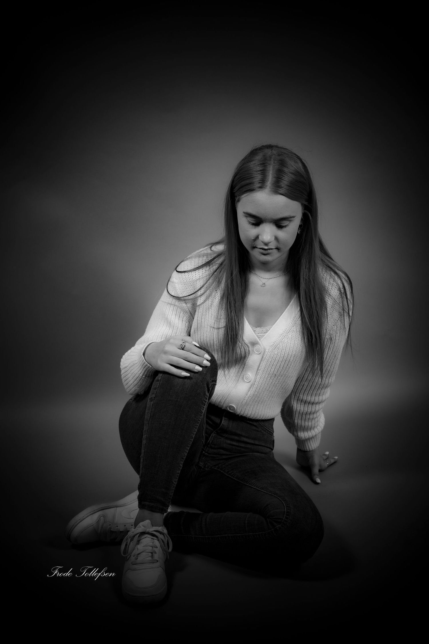 Black and white studio portrait of a young woman sitting on the floor in a white sweater and dark jeans.
