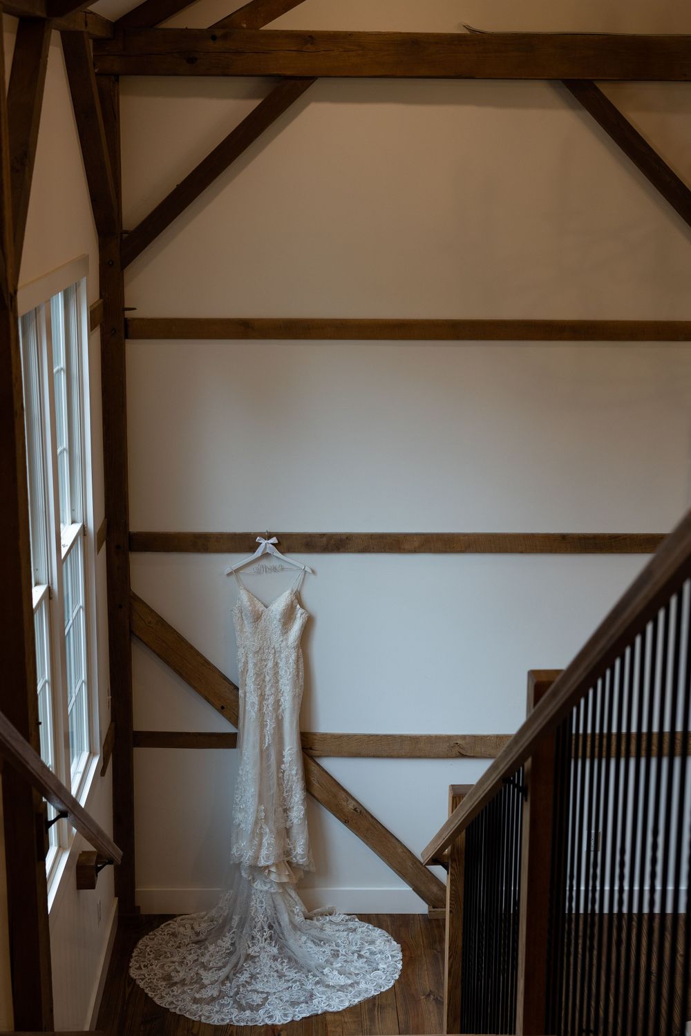 Elegant lace wedding dress hanging against rustic wooden beams in natural light.