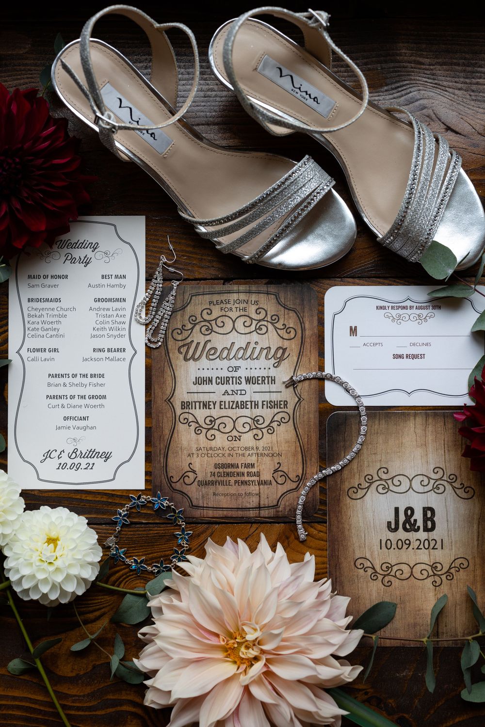 Wedding invitation suite with silver shoes and pink dahlias on dark wooden surface.
