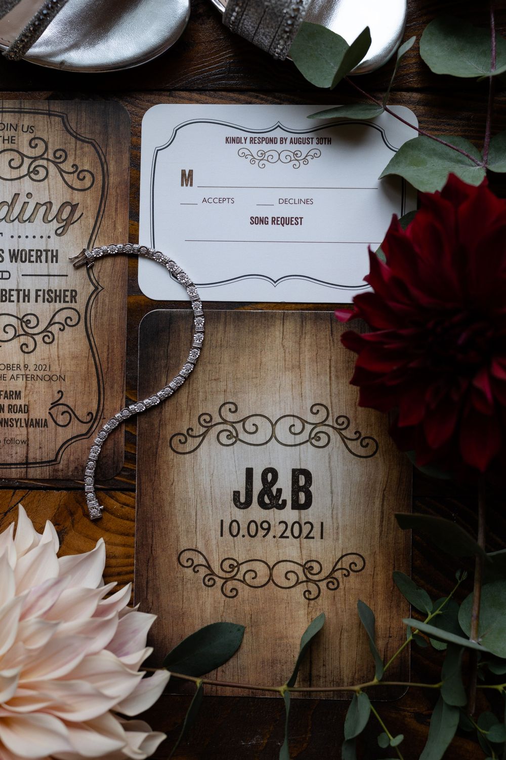 Wooden wedding invitation with initials J&B and date 10.09.2021 surrounded by red and pink flowers and greenery.