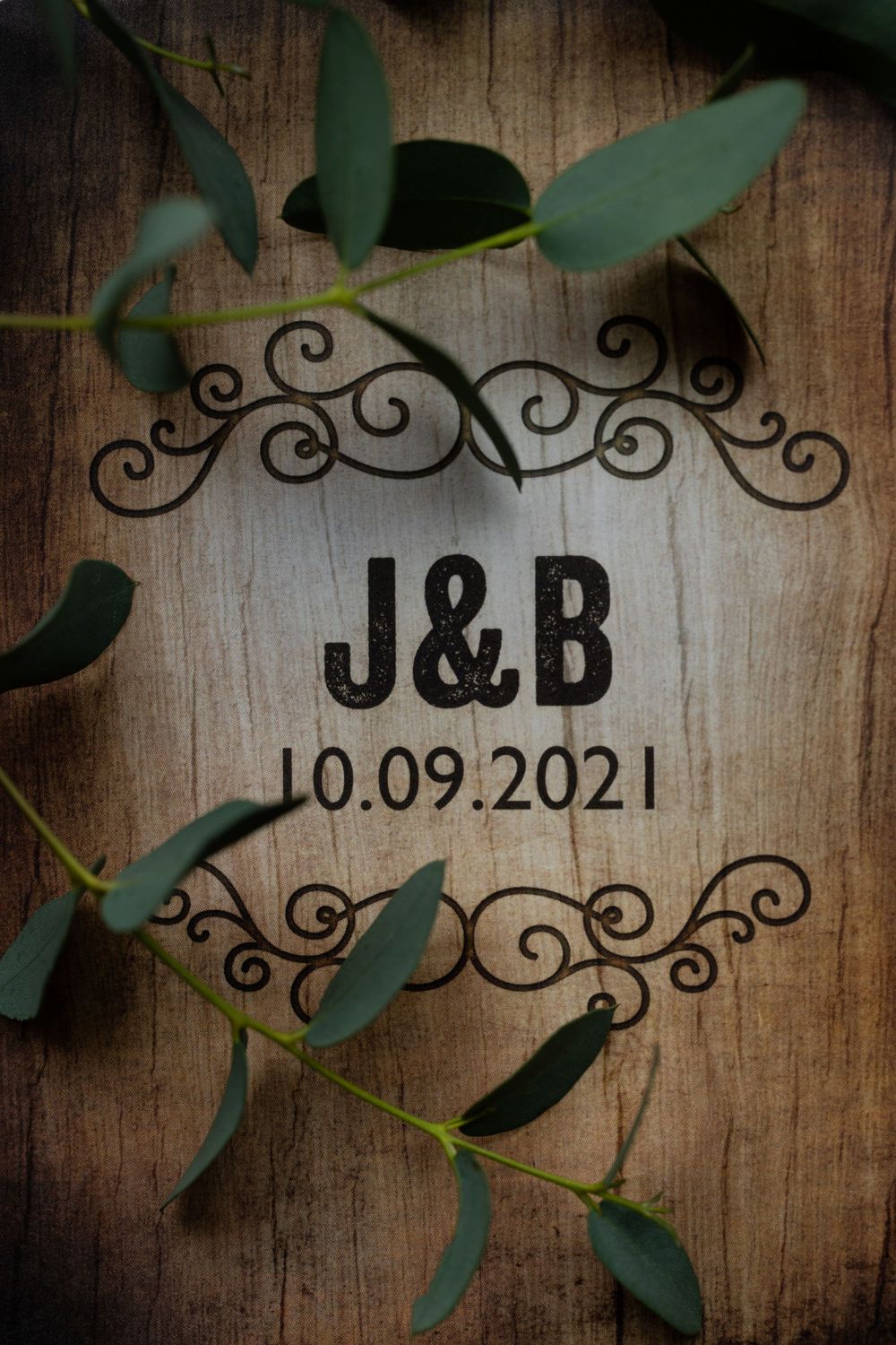 Wooden wedding sign with J&B monogram and date framed by decorative scrollwork and eucalyptus leaves.