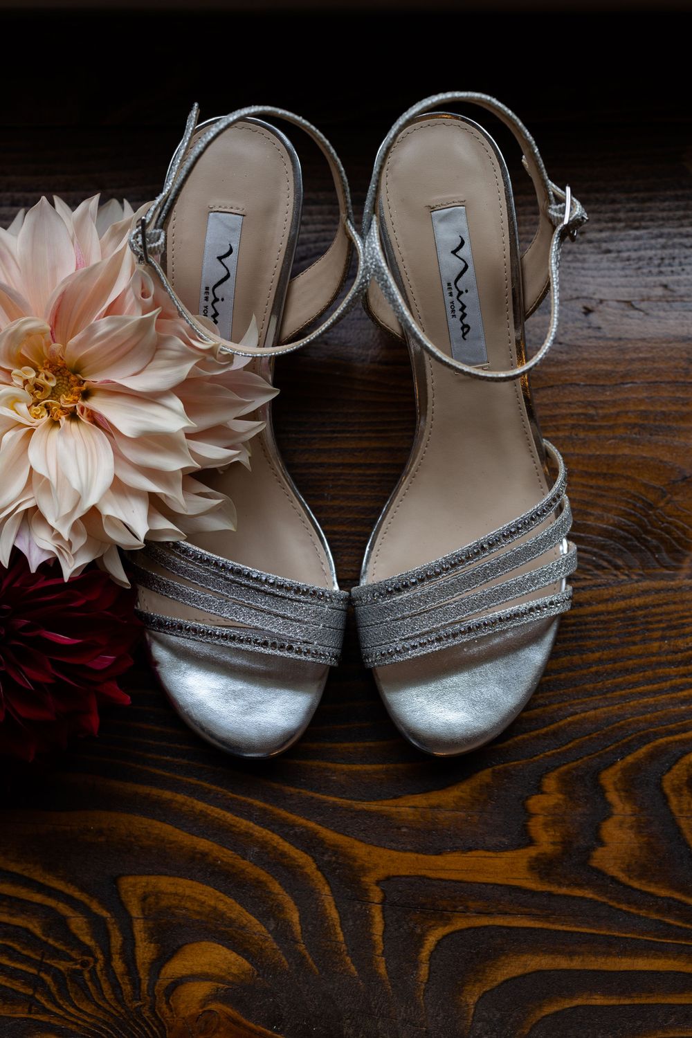 Silver strappy wedding sandals next to pink and burgundy flowers on wooden surface.