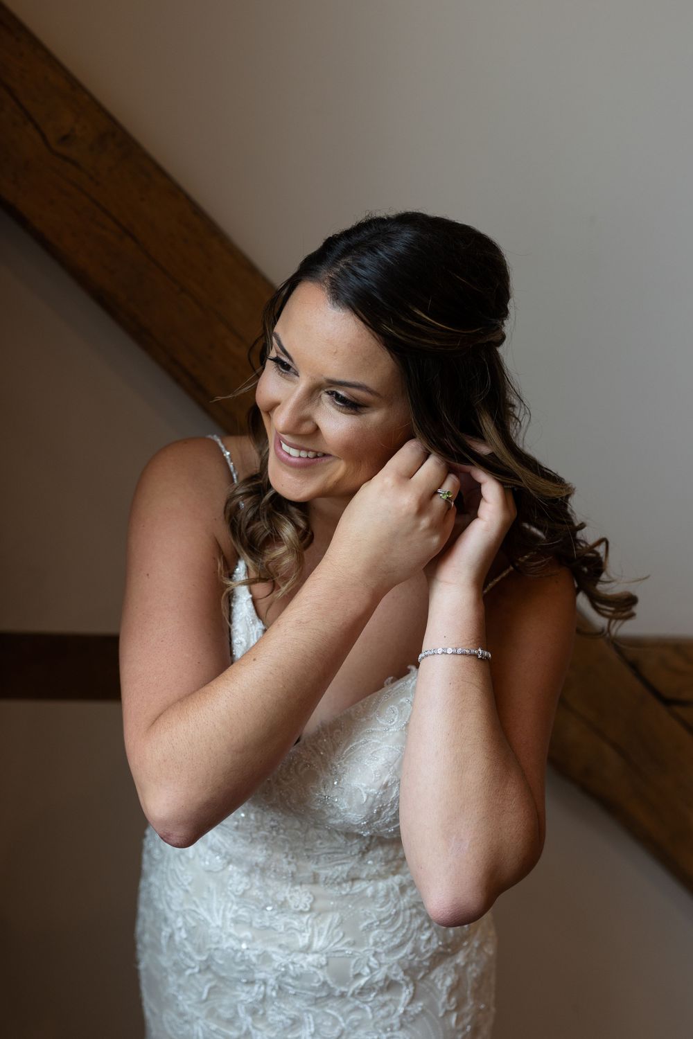 Bride in a white lace dress adjusts her earrings while smiling warmly in a rustic indoor setting.