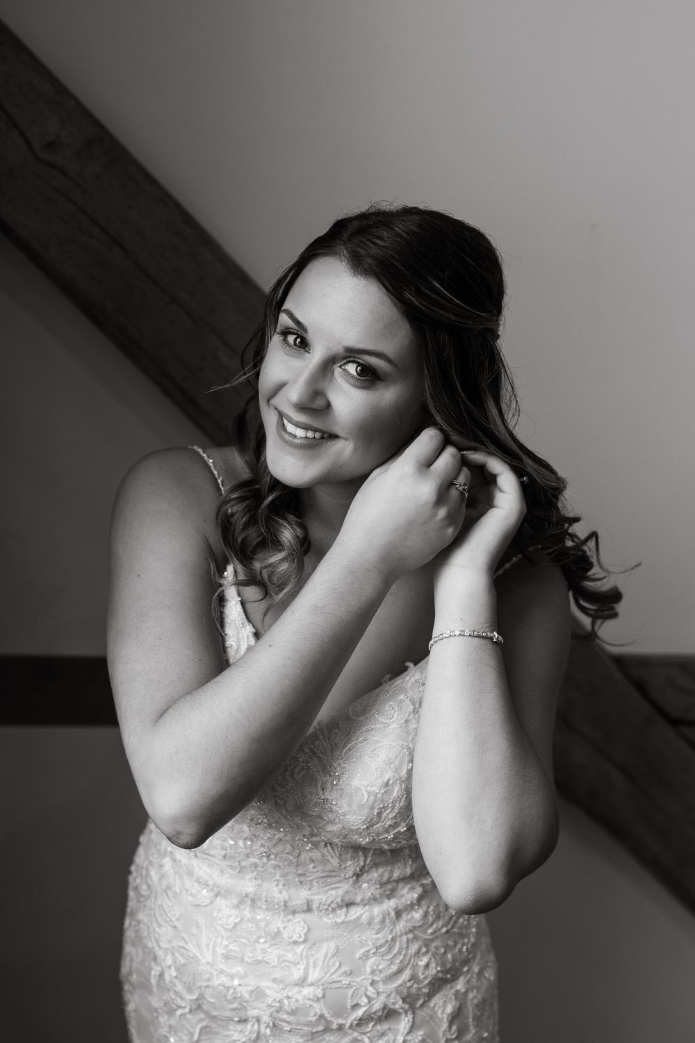 Black and white photo of a bride in lace dress touching her earrings with a joyful expression.