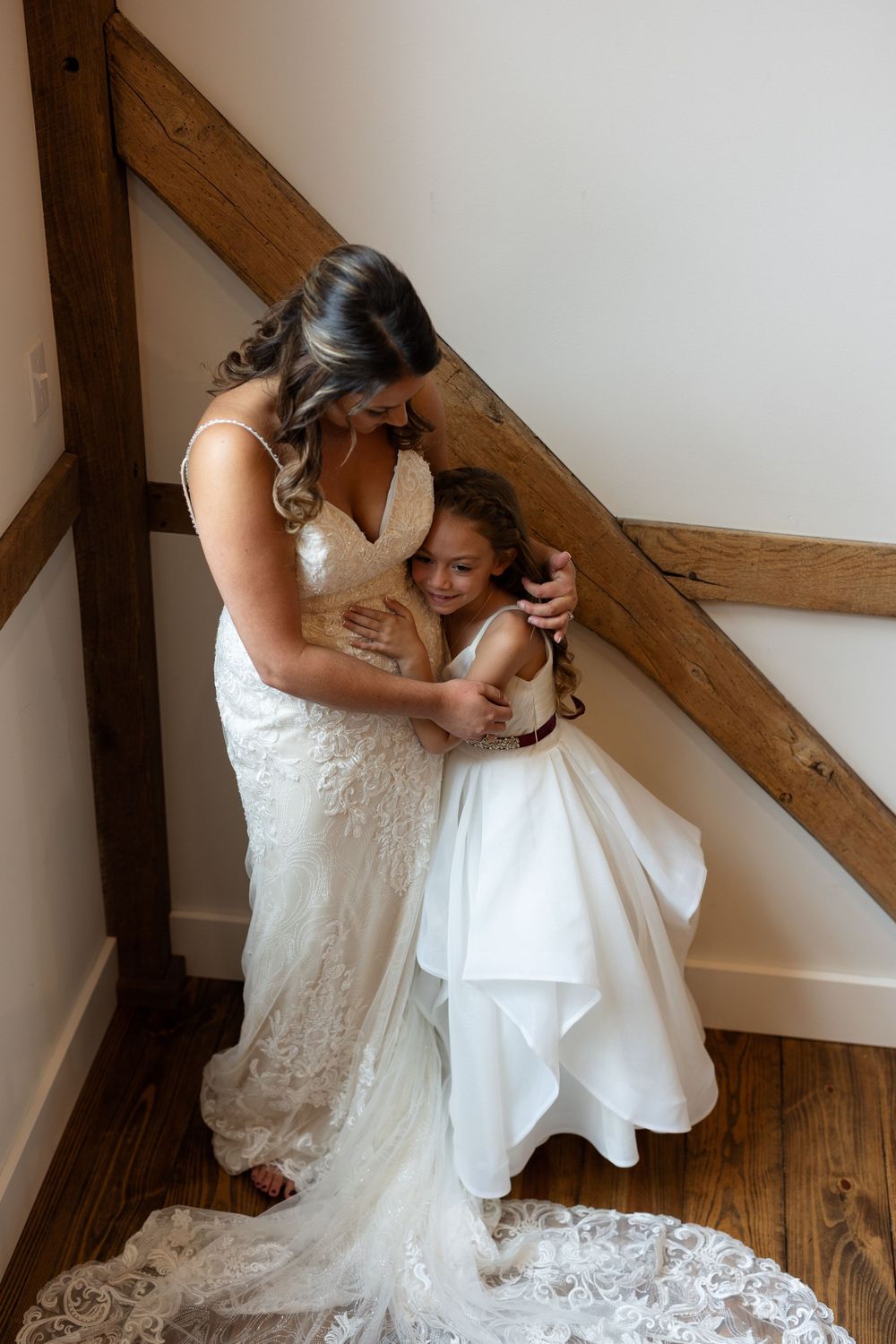 Touching moment between bride in lace gown and young flower girl in white dress near wooden beam.