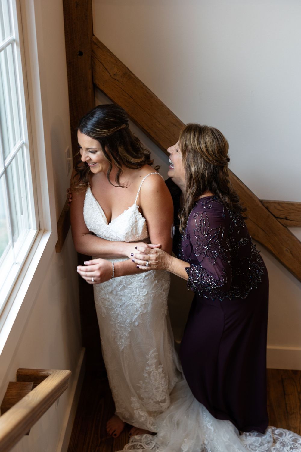 Two women share laughter while standing by a window in wedding attire.