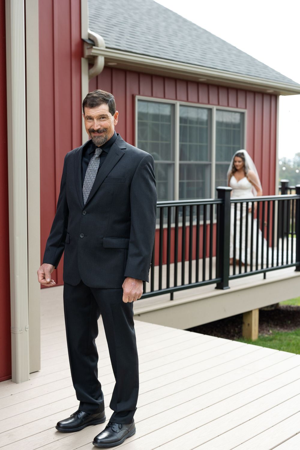 Man in dark suit stands on wooden deck with red barn-style building in background.