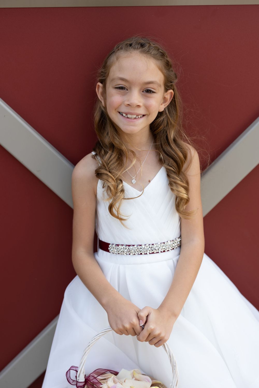 Young flower girl in white dress with sparkly belt smiling in front of red barn door.