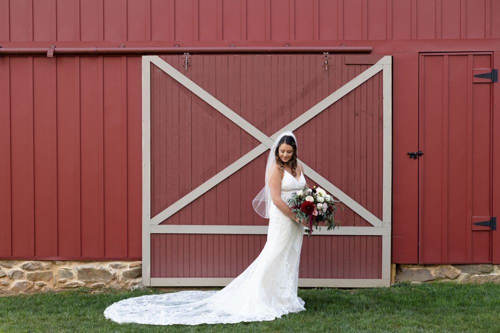 Bride in lace wedding gown poses against red barn door with white X pattern.