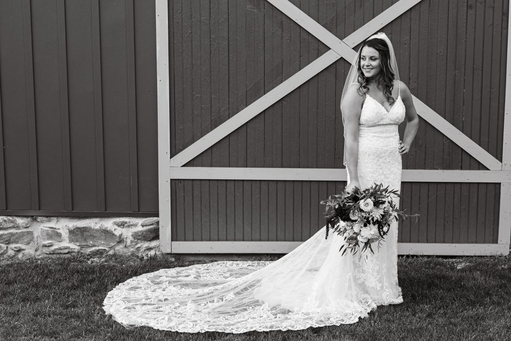 Black and white portrait of bride with long train against rustic barn backdrop.